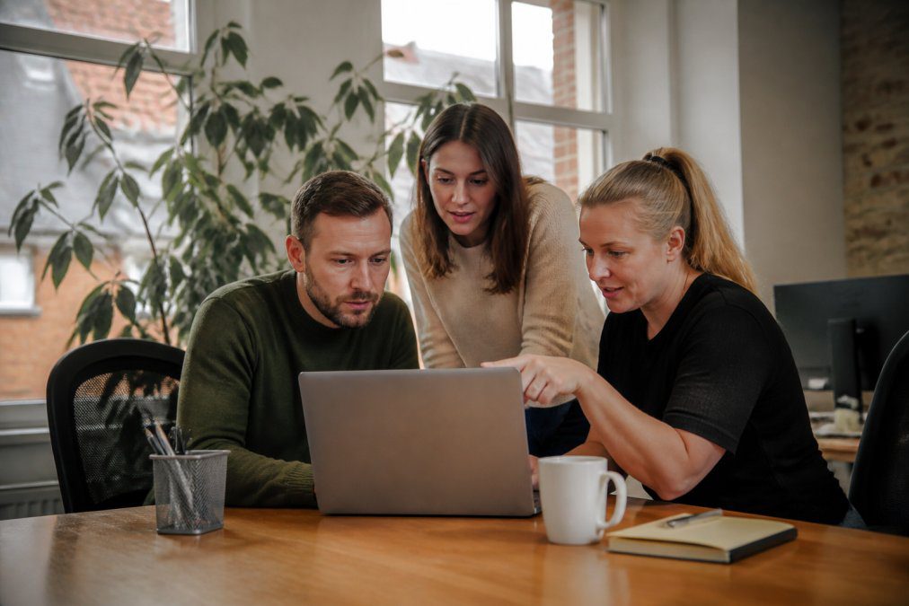 A small office team in Norfolk gathered around a laptop screen, one colleague reviewing remote access settings while another points at the screen, natural daylight through office windows